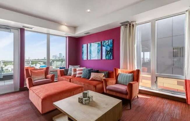 A vibrant resident lounge here at Blue and Lime showcasing a cozy seating area with a rust-toned sectional sofa, matching accent chairs, and a square wooden coffee table set on textured carpeting. Floor-to-ceiling windows fill the space with natural light, while a bold magenta accent wall, modern artwork, and soft window curtains add color and warmth.