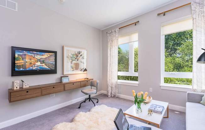 The Residences at Crosspoint living room corner features a wall-mounted flat-screen TV, abstract artwork above a floating wood console, a sheepskin rug on the floor, and a white coffee table holding a vase of yellow tulips.