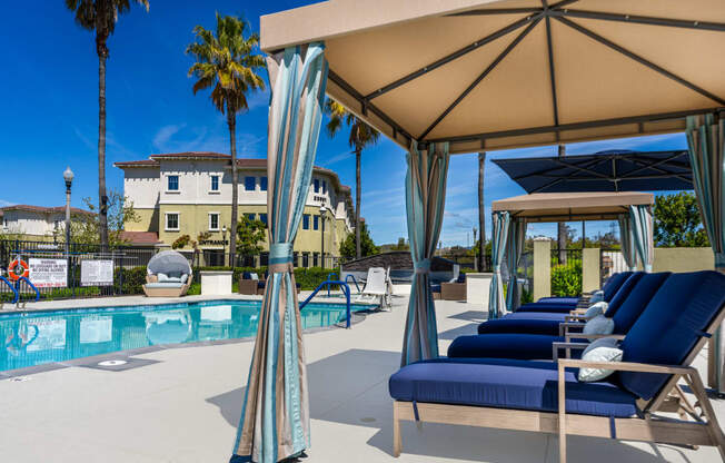 a pool with blue lounge chairs and a tan umbrella next to a pool