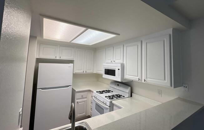 A clean, modern kitchen featuring white cabinets, a white refrigerator, a microwave above the stove, and a gas range. The countertop is light-colored, and the space is well-lit with ceiling lights. The kitchen has a sleek and inviting appearance, suitable for cooking and dining.