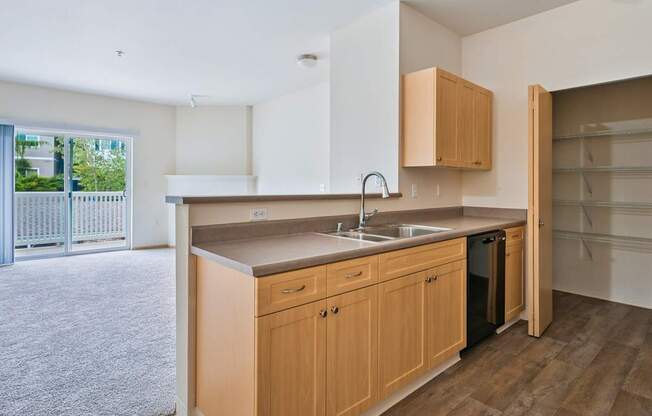 Soho kitchen with wooden cabinets, a pantry closet, and the living room in the background at Abbey Rowe Apartments in Olympia, WA