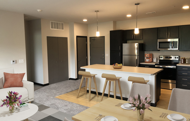 A modern kitchen and dining area featuring a central island with stools, stainless steel appliances, a dining table set with flowers, and a cozy living space with a white sofa. The decor is minimalistic with neutral tones and warm lighting, creating a welcoming atmosphere.