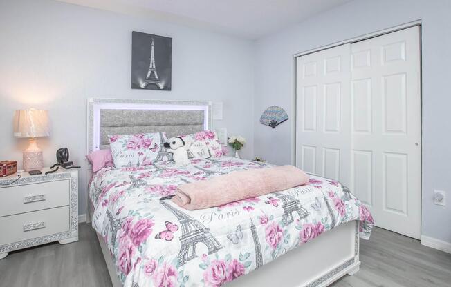 A cozy bedroom featuring a bed with a pink floral duvet cover and decorative pillows. There's a bedside table with a lamp, a small panda plush toy on the bed, and light blue walls. A closet door is visible in the background. Decor includes an Eiffel Tower picture above the bed for a Parisian touch.