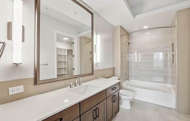 A modern bathroom from the C1 floorplan with a large mirror, white counter with wooden cabinets, sink, and bathtub at Alastair at Aria Village Apartments in Sandy Springs, GA