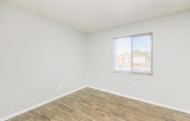 Bright, empty room with light gray walls and a large window allowing natural light to enter. The floor is covered in wooden planks, giving the space a warm feel. No furniture is present, highlighting the spaciousness of the area. The window faces an outside view with light filtering through.