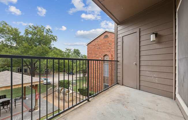 A balcony with a black railing overlooks a courtyard with a brick building in the background.