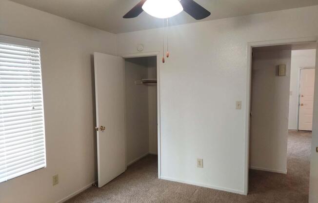 A well-lit, empty room featuring beige carpet, white walls, and a ceiling fan. There are two doors; one leads to a closet and the other to another room. A window with white blinds allows natural light to enter. The overall space appears clean and ready for furnishing.