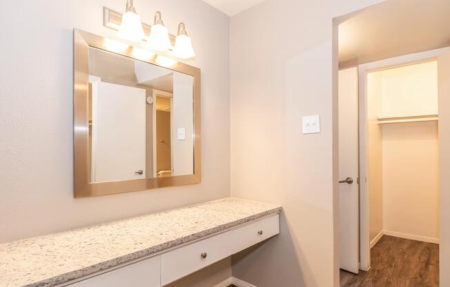 A well-lit vanity area featuring a large mirror with three light fixtures above, a countertop made of granite or a similar material, and a white cabinet below. To the right, there's an open door leading to a small closet with a hanging rod, set against a neutral wall color and dark laminate flooring.