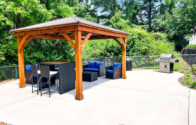 A wooden gazebo with a table and chairs is surrounded by trees.