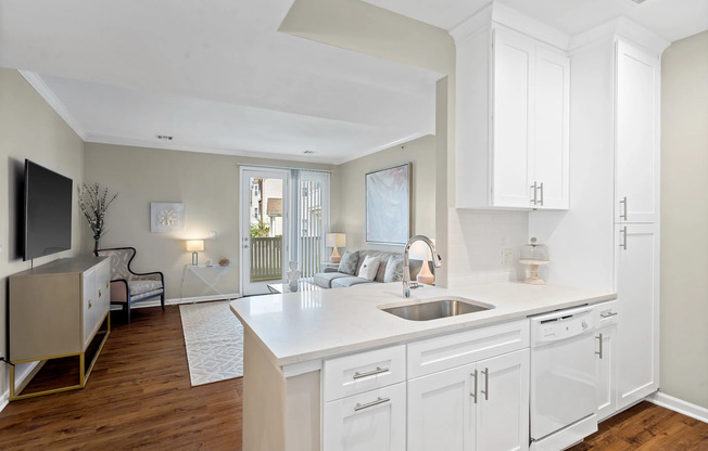 Bright, open-concept apartment interior featuring white shaker kitchen cabinets, stainless steel sink, modern faucet, white quartz countertops, wood-style flooring, and a cozy living room with natural light and private patio access at Kelly’s Ridge Apartments in Columbia, Missouri.
