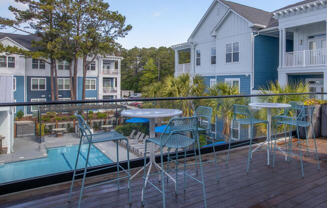 A balcony view featuring two white tables and blue chairs overlooking a swimming pool surrounded by palm trees, with modern apartment buildings in the background. The scene is bright and inviting, showcasing a relaxed outdoor space.
