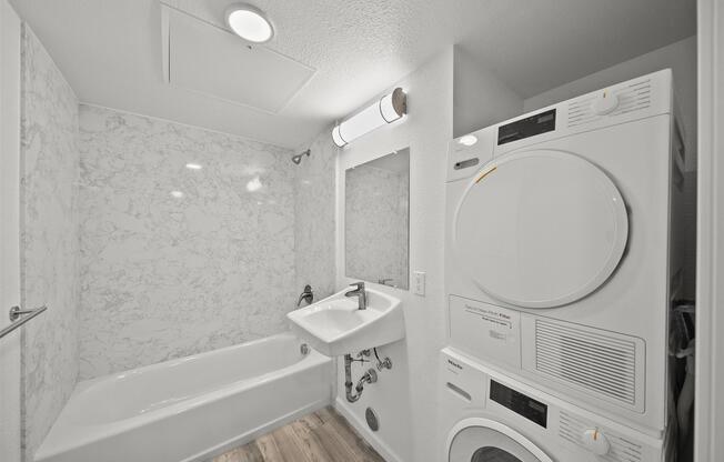 A modern bathroom featuring a white tub with a marble backsplash, a sink, a large mirror, and a stacked washer and dryer unit. The space is well-lit with a ceiling light and has a minimalist design with light wood flooring.