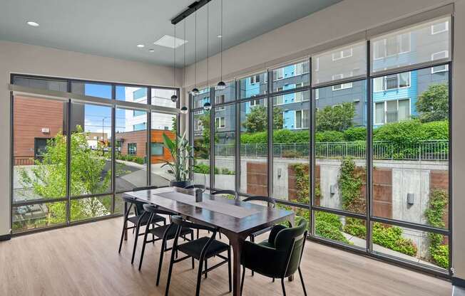 A dining table with chairs is set up in a room with a view of a building and greenery outside.