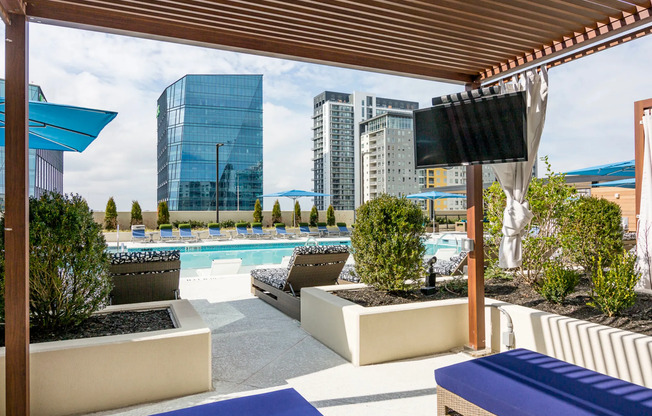 A wooden cabana space with two upholstered lounge chairs overlooking the pool and city skyline, enclosed by a raised landscaping bed with bushes.