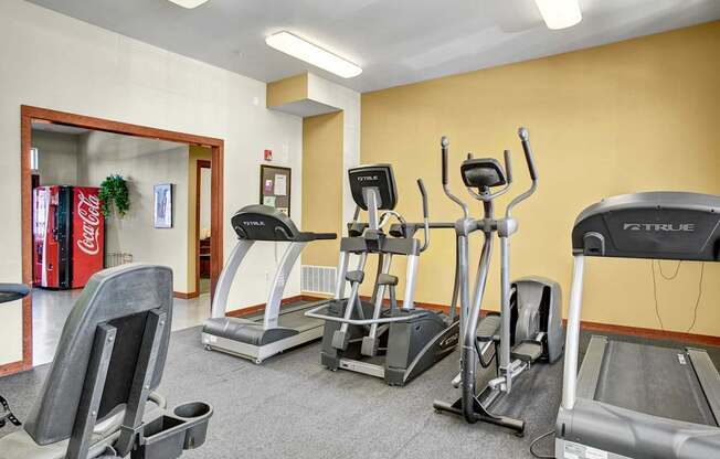 A well equipped fitness center here at Allegro featuring multiple cardio machines including treadmills and elliptical trainers arranged on soft flooring, with bright overhead lighting, warm neutral walls, and an adjacent lounge area visible through an open doorway, creating a convenient and inviting space for daily workouts.