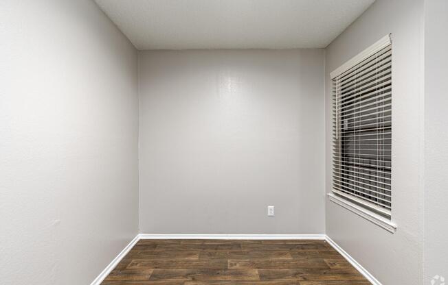 Empty room with light gray walls and a hardwood floor. A single window with white blinds is located on one wall, creating a bright, minimalist space. The corner of the room is visible, emphasizing its spaciousness and simplicity. No furniture or decorations are present.