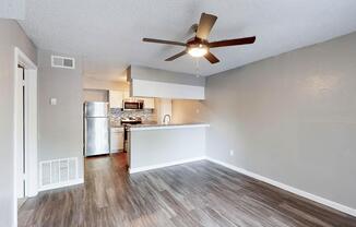 A spacious living area featuring grey walls and a ceiling fan. There is a view of a modern kitchen with a stainless steel refrigerator and a stylish backsplash. The flooring is a light wood finish, creating a welcoming atmosphere. Natural light fills the room, enhancing the open layout.