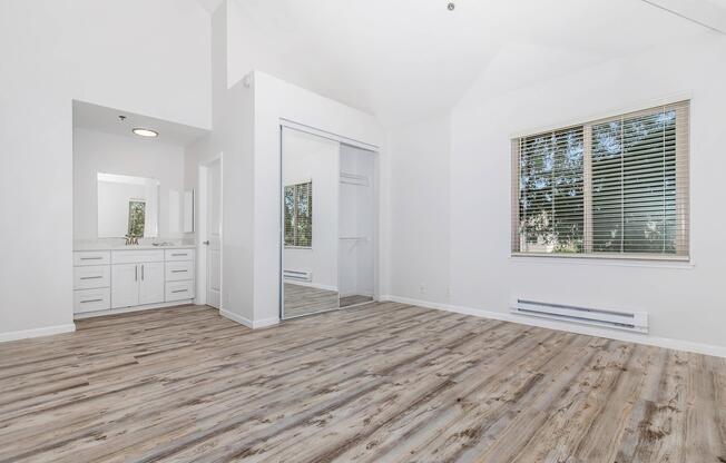 A spacious, bright room with white walls and a high ceiling. It features a large window with blinds, showcasing natural light. On one side, there's a modern bathroom vanity with a mirror, and on the opposite wall, a sliding glass closet door. The floor is a warm, light-colored hardwood.