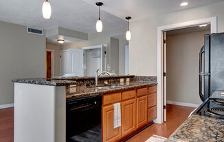 A kitchen here at Bridges with a granite breakfast bar and island sink, warm wood cabinets, black appliances, pendant lighting, and open sightlines to the living area and nearby rooms.