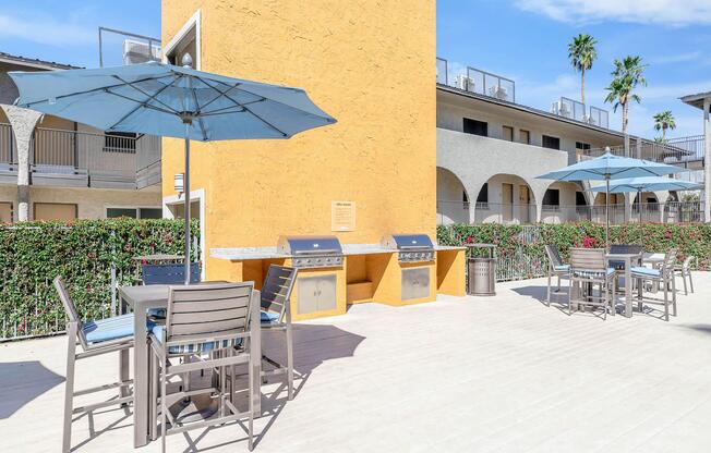 Outdoor grill area featuring two barbecue stations under blue umbrellas, surrounded by patio seating. The setting includes palm trees and modern apartment buildings in the background, with a sunny sky above and greenery along the edges, creating a relaxed and inviting atmosphere.