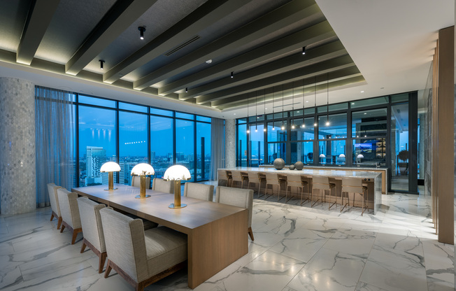 Resident lounge with an 8-person table for dining or work, a work area and wet bar with 7 chairs along a marble desk, and two full-width floor-to-ceiling windows with a city view. The floors, walls, and columns are marble-clad.