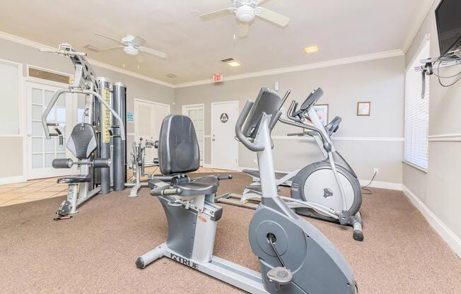 An indoor gym featuring exercise equipment including an elliptical machine, a stationary bike, a multi-station weight machine, and a ceiling fan. The walls are painted in light colors, and there is a television mounted on one wall. The flooring is covered with carpet, creating a comfortable workout environment.