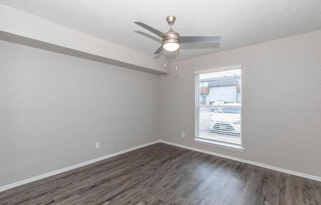 Empty room with light gray walls and dark wood-style laminate flooring. A ceiling fan with a light is visible, and there is a window with white blinds letting in natural light. The overall atmosphere is bright and spacious.