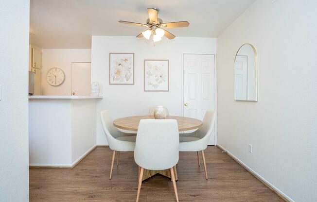 Modern dining area featuring a round wooden table with light-colored upholstered chairs. A ceiling fan is overhead, and soft white walls create a bright atmosphere. Two framed floral artworks hang on the wall, complemented by a mirror. The space is open with a view into the adjacent kitchen area.