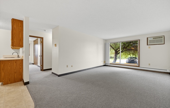 A spacious living room with a grey carpet and a window overlooking a tree.