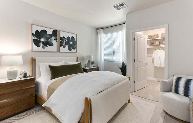 A serene bedroom here at Reflect at Dobson Ranch featuring a cozy upholstered bed with crisp white bedding and layered accent pillows, matching wood nightstands with modern lamps, soft neutral walls, and a window with blinds and sheer curtains, with an open walk-in closet visible and plush carpeting creating a calm, inviting atmosphere.