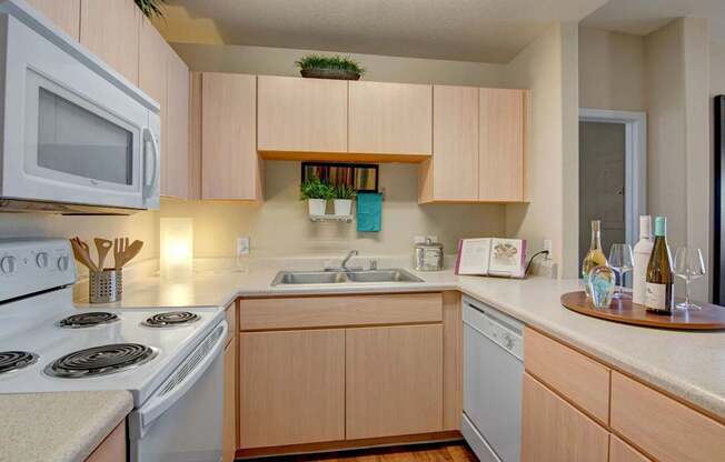 A warm and inviting kitchen here at Pala Mesa featuring light wood cabinetry, expansive countertops, and a double-basin sink with sleek faucet. A full appliance package including a range, built-in microwave, and dishwasher enhances everyday convenience, while wood-style flooring and soft overhead lighting create a clean, comfortable cooking space.