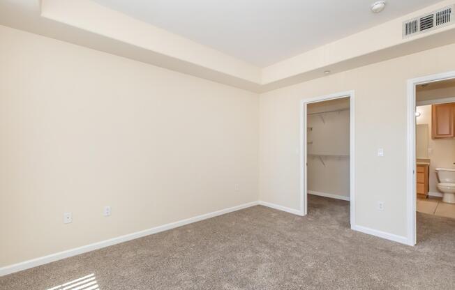 A spacious, empty room with light beige walls and carpet. There is a closet on one side and a doorway leading to a bathroom on the other. Natural light enters through a window, enhancing the bright and open feel of the space.