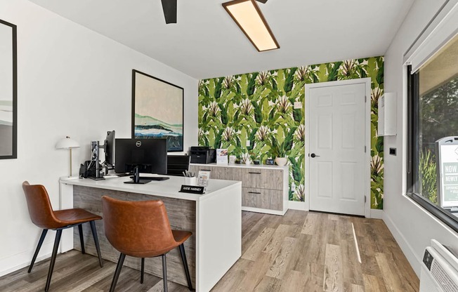 A room with a desk and chairs in front of a window with a view of a green plant.