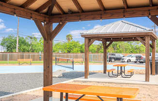 A wooden pavilion with picnic tables is situated in a park with a playground in the background.