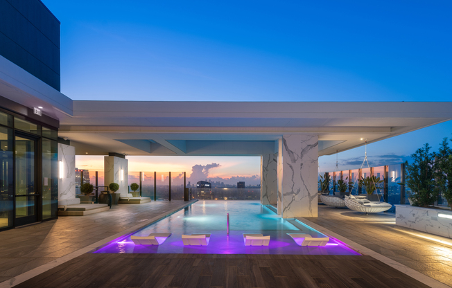 An outdoor lap pool deck with a wide overhang shading part of the pool. There are purple underwater lights shining on 4 in-pool lounge chairs, marble-clad landscaping boxes with large bushes, and a sunset view of the downtown Houston skyline.