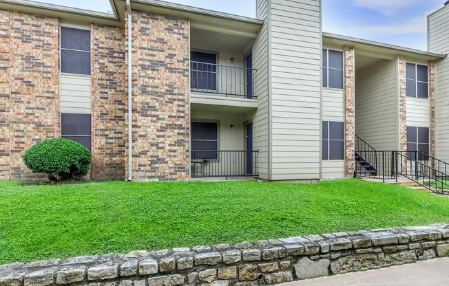 A multi-unit apartment building featuring a combination of brick and siding exterior, with two levels, grassy areas, and a stone wall in the foreground.