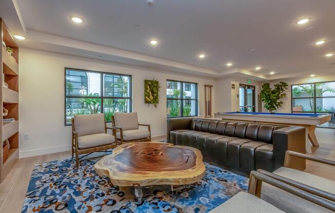 A modern lounge area featuring a wooden coffee table, comfortable seating with gray chairs and a dark leather sofa. The space has large windows allowing natural light, complemented by indoor plants and a pool table, creating a welcoming atmosphere for relaxation and socializing.