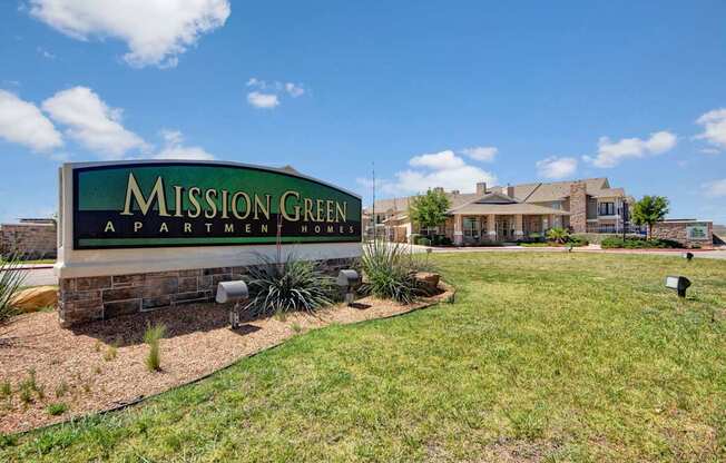A welcoming entrance sign here at Mission Green displaying the community name set on a stone base, surrounded by manicured landscaping, decorative grasses, and a wide green lawn, with apartment buildings, walkways, and a bright blue sky with scattered clouds in the background.