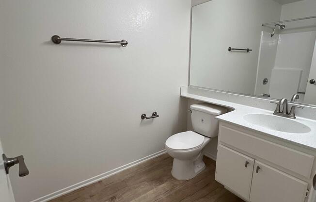 A clean, minimalist bathroom featuring a white toilet, a single sink with a countertop, a large mirror, and a towel rack. The walls are painted light, and the floor is made of wood. The space is well-lit and uncluttered, with basic fixtures and no decorative elements.