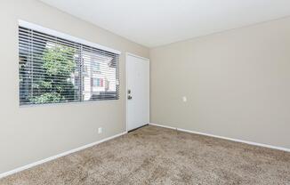 A well-lit, empty room with light beige walls and soft, brown carpeting. There is a white door leading outside and a large window with blinds, allowing natural light to enter. The space is uncluttered, offering a sense of openness and potential for interior design.