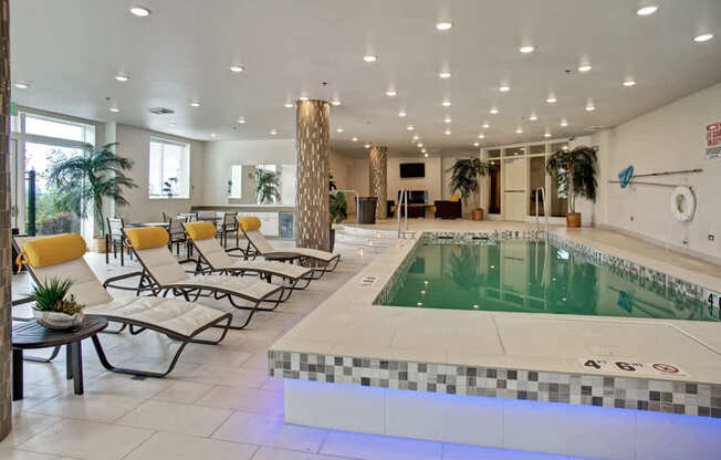 A resort-style indoor pool here at Heritage Hills featuring a rectangular swimming pool with clear water, a tiled pool deck, and stainless handrails along the edge. The space includes cushioned lounge chairs with yellow accent pillows, side tables, tall patterned columns, potted greenery, and bright recessed ceiling lighting creating a clean, inviting atmosphere.