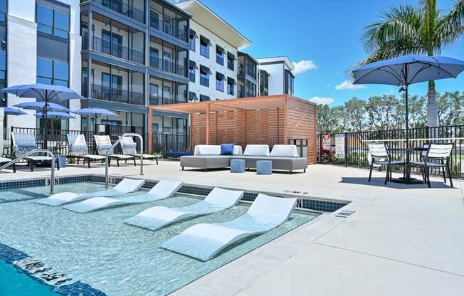 a pool with lounge chairs and umbrellas in front of an apartment building