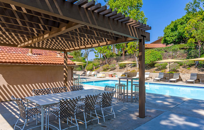 A table and chairs are set up under a pergola next to a pool.