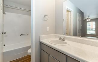 A clean and modern bathroom featuring a bathtub, a sink with a countertop, and a mirror. The walls are painted in neutral tones, and there is a towel ring on the wall. The door leads to another room with natural light coming through a window, enhancing the spacious feel of the area.