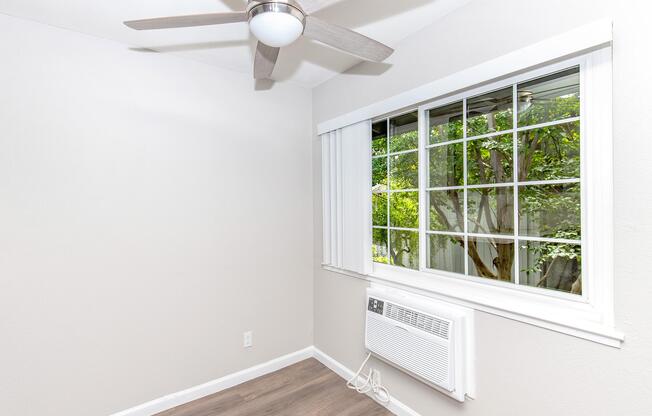 A well-lit room featuring a light-colored wall, a ceiling fan with a light fixture, a large window with white blinds revealing greenery outside, and a wall-mounted air conditioning unit below the window. The flooring is a light wood laminate, contributing to a clean and modern aesthetic.