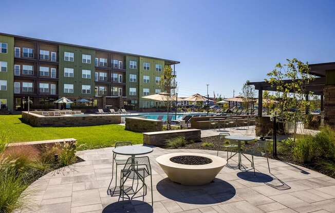 A patio with a fire pit and chairs is surrounded by a grassy area and a building.