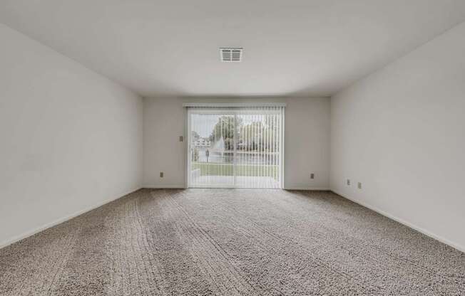 A room with a carpeted floor and a view of a fence and trees outside through a sliding glass door.