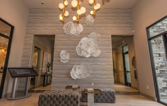 A stylish lobby area featuring a textured wall with large, decorative white floral wall art. The space is well-lit by a modern chandelier with multiple bulbs. Two tufted benches and a sleek information stand are also present, creating an inviting atmosphere.