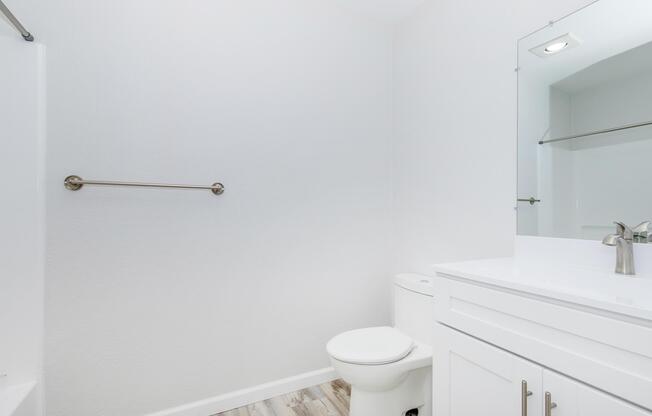 A clean, modern bathroom featuring a white toilet and a sleek vanity with a sink. The walls are painted a light color, and there is a large mirror above the sink. A towel bar is mounted on the wall, and the floor has a light wooden texture, creating a bright and spacious atmosphere.