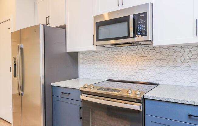 A modern kitchen featuring a stainless steel refrigerator, a built-in microwave above a smooth-top stove, and blue cabinetry with white countertops. The backsplash consists of geometric patterned tiles, creating a stylish and contemporary look.